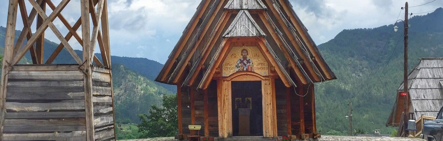 Una iglesia de madera con un campanario y una cruz en la parte superior, rodeada de montañas y un cielo nublado.