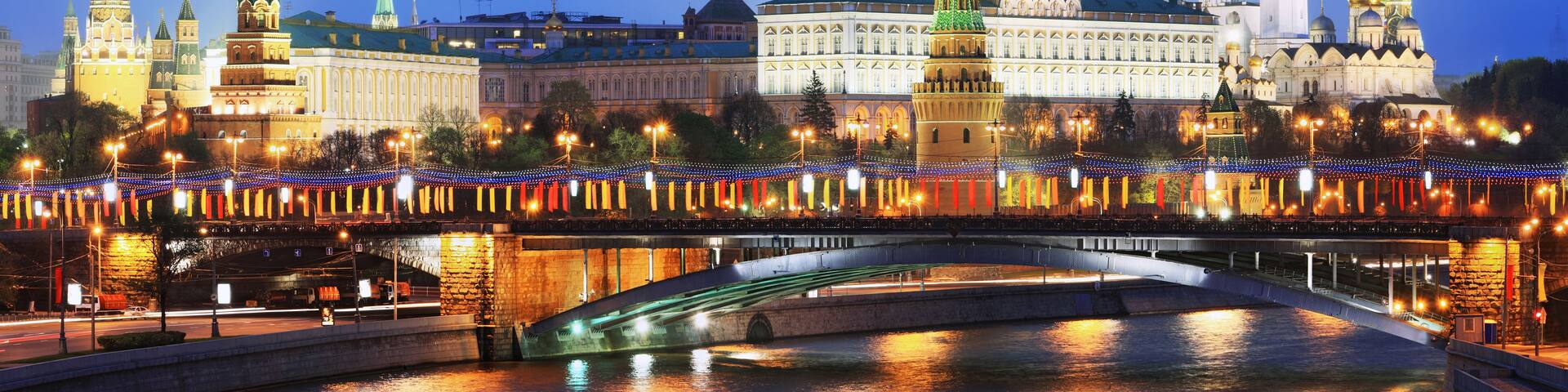 A river with a bridge, illuminated buildings, and a prominent tower with a flag.