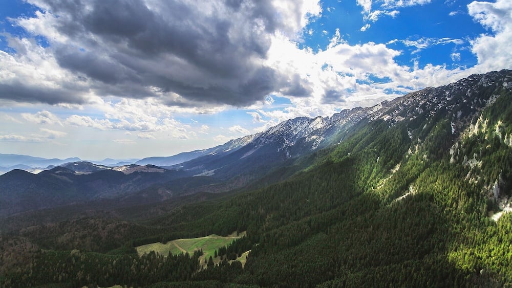 A mountainous landscape with a valley, dense forests, and a clear sky with clouds.