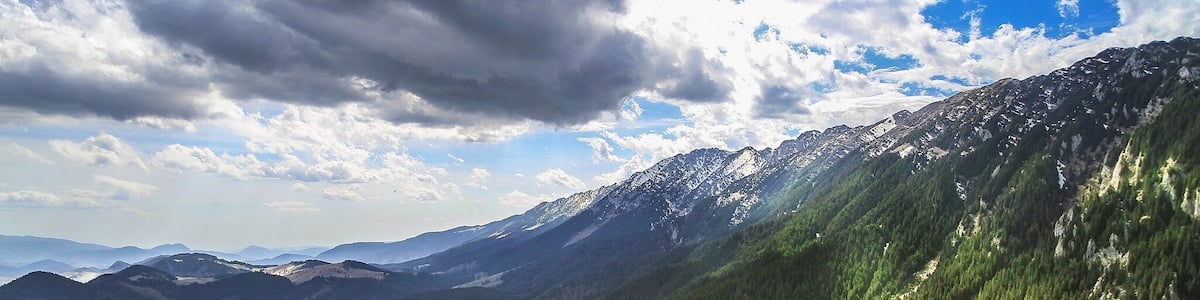 A mountainous landscape with a valley, dense forests, and a clear sky with clouds.