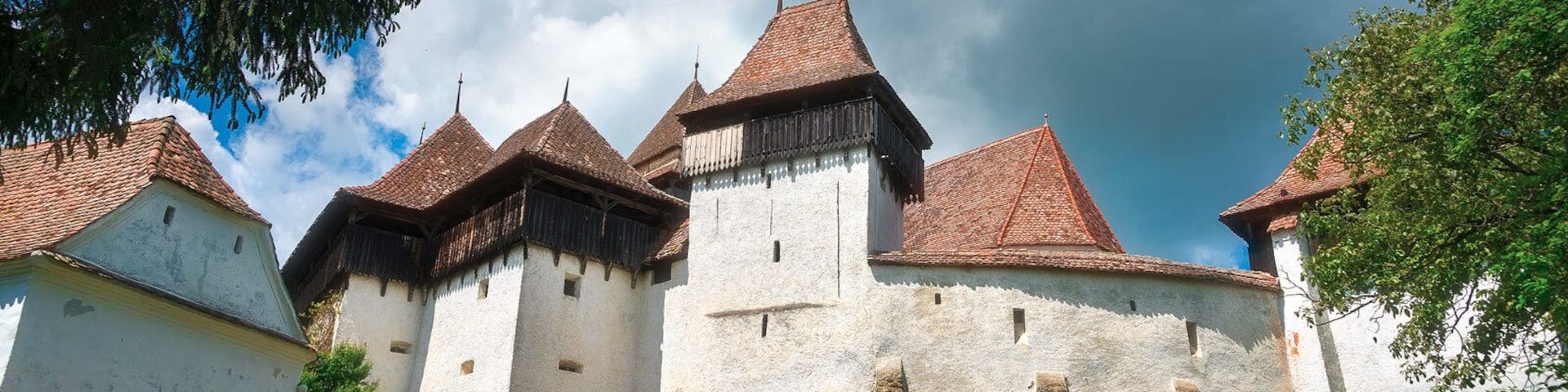 A historic castle with multiple towers and a white facade, surrounded by lush greenery and a partly cloudy sky.