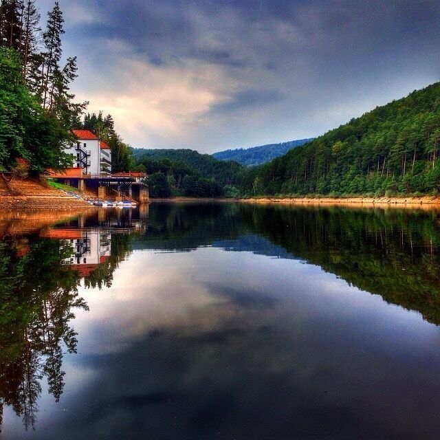 A calm lake with a house on the left and a forested hillside on the right.