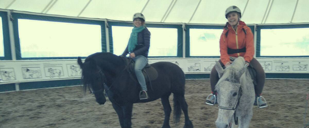 Two people riding horses inside a tent with a sandy floor and windows.