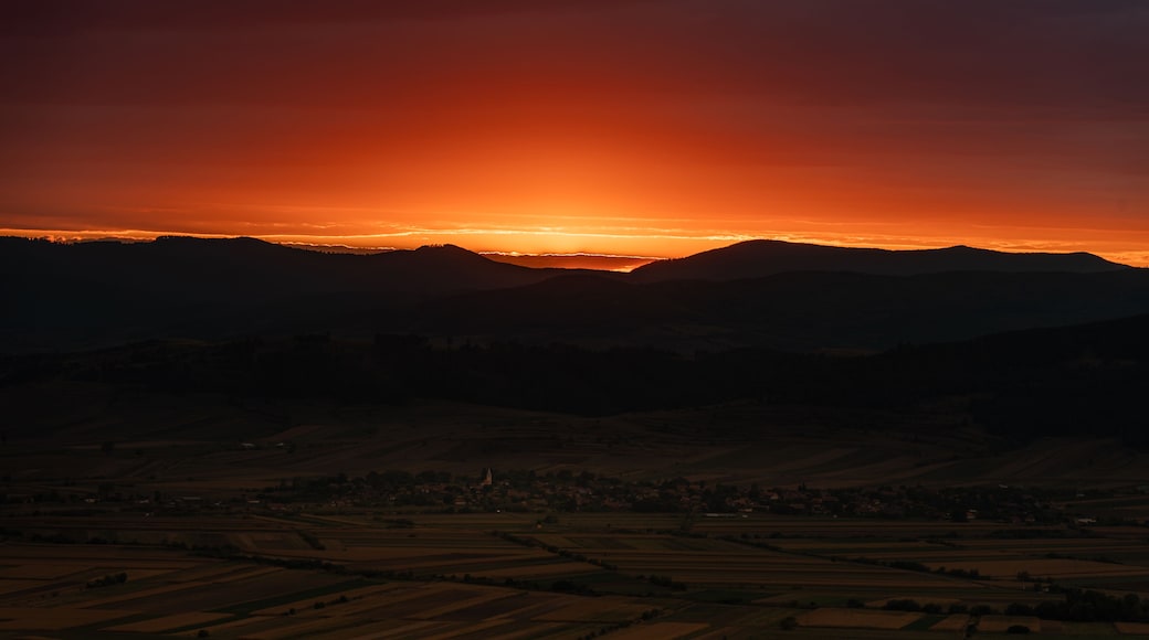 View of a vibrant sunset casting a warm orange glow over the rolling hills and agricultural fields surrounding the village of Mereni, Covasna County, Romania.