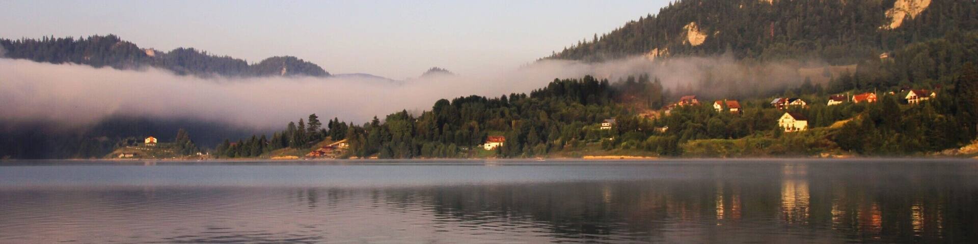 A calm lake with reflections of houses and mountains, surrounded by forested hills and mist.