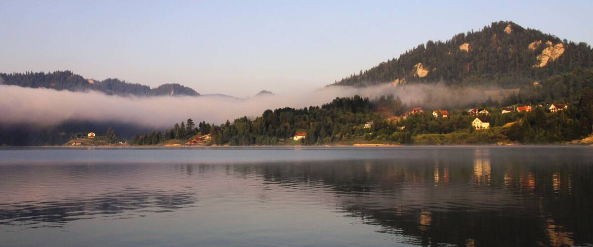 A calm lake with reflections of houses and mountains, surrounded by forested hills and mist.
