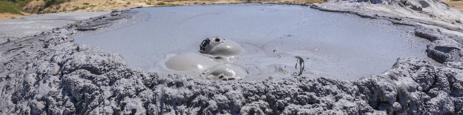 A geothermal area with bubbling mud pools, rocky terrain, and green hills in the background.