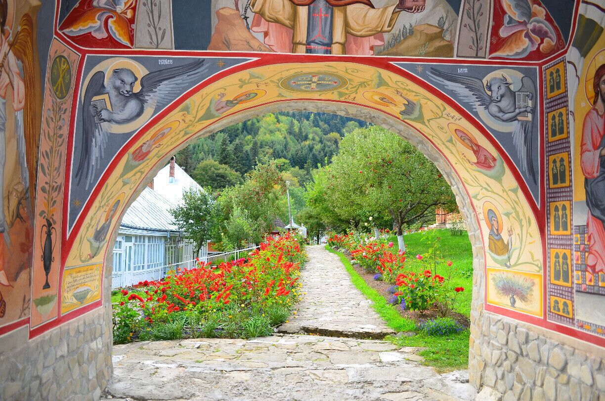 A stone pathway leading through an ornate archway with colorful frescoes, surrounded by vibrant flower beds and a mountainous backdrop.