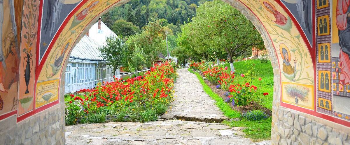 A stone pathway leading through an ornate archway with colorful frescoes, surrounded by vibrant flower beds and a mountainous backdrop.