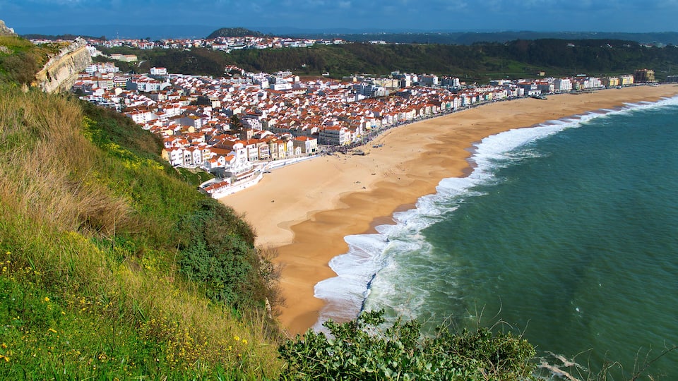 A coastal town with a sandy beach, a rocky cliff, and a dense cluster of red-roofed buildings.