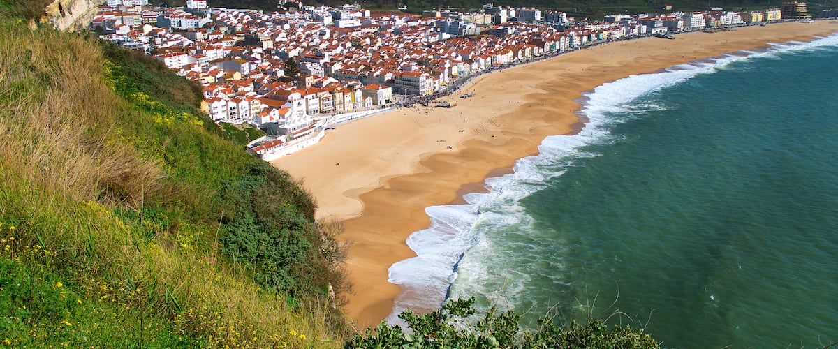A coastal town with a sandy beach, a rocky cliff, and a dense cluster of red-roofed buildings.