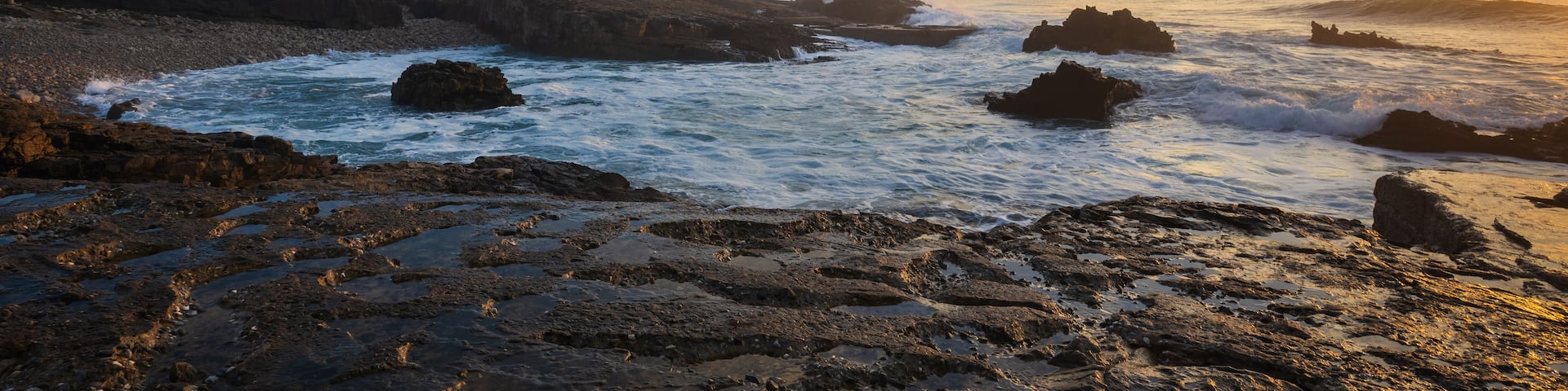 A lighthouse, rocky shoreline, and waves crashing against rocks at sunset.