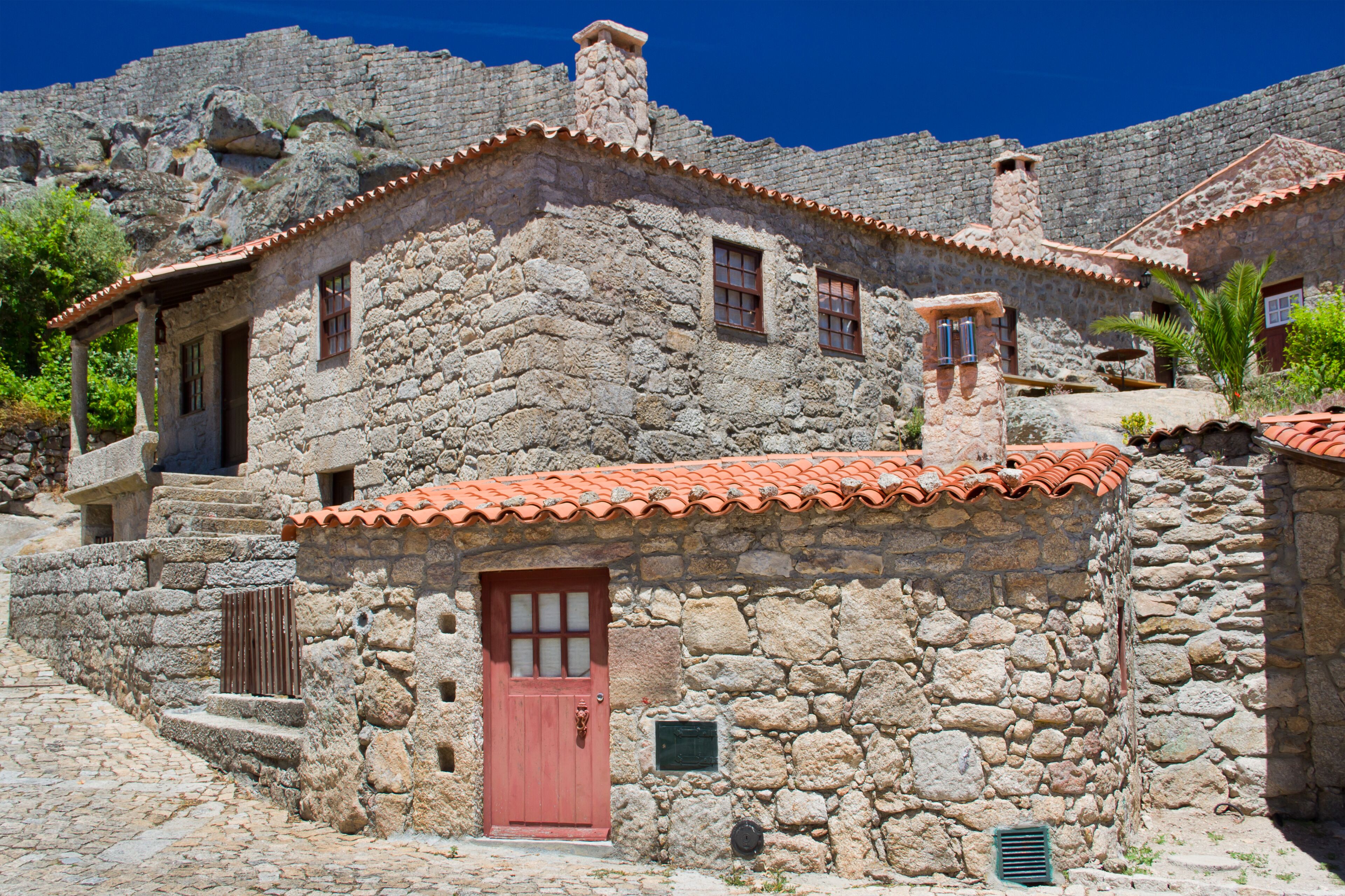 A stone building with a red door and windows, surrounded by cobblestone streets and greenery.