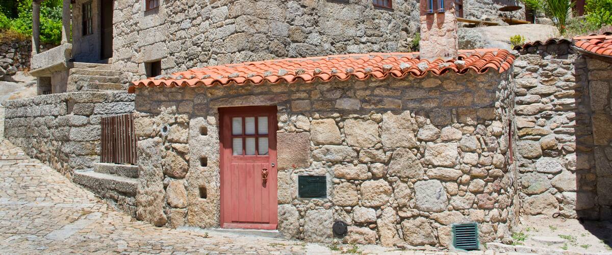 A stone building with a red door and windows, surrounded by cobblestone streets and greenery.