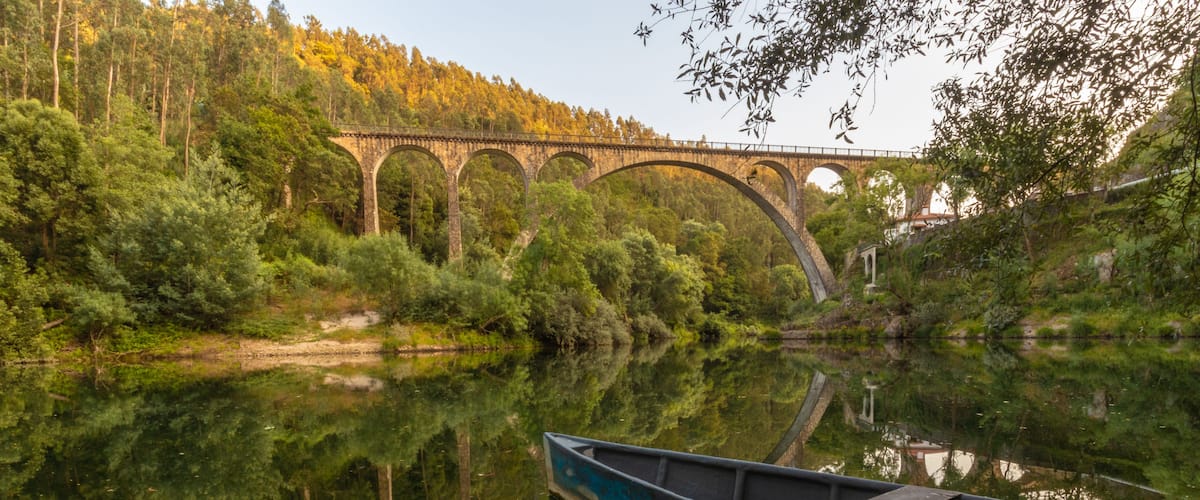A calm river with a wooden boat, dense greenery, and a stone bridge in the background.