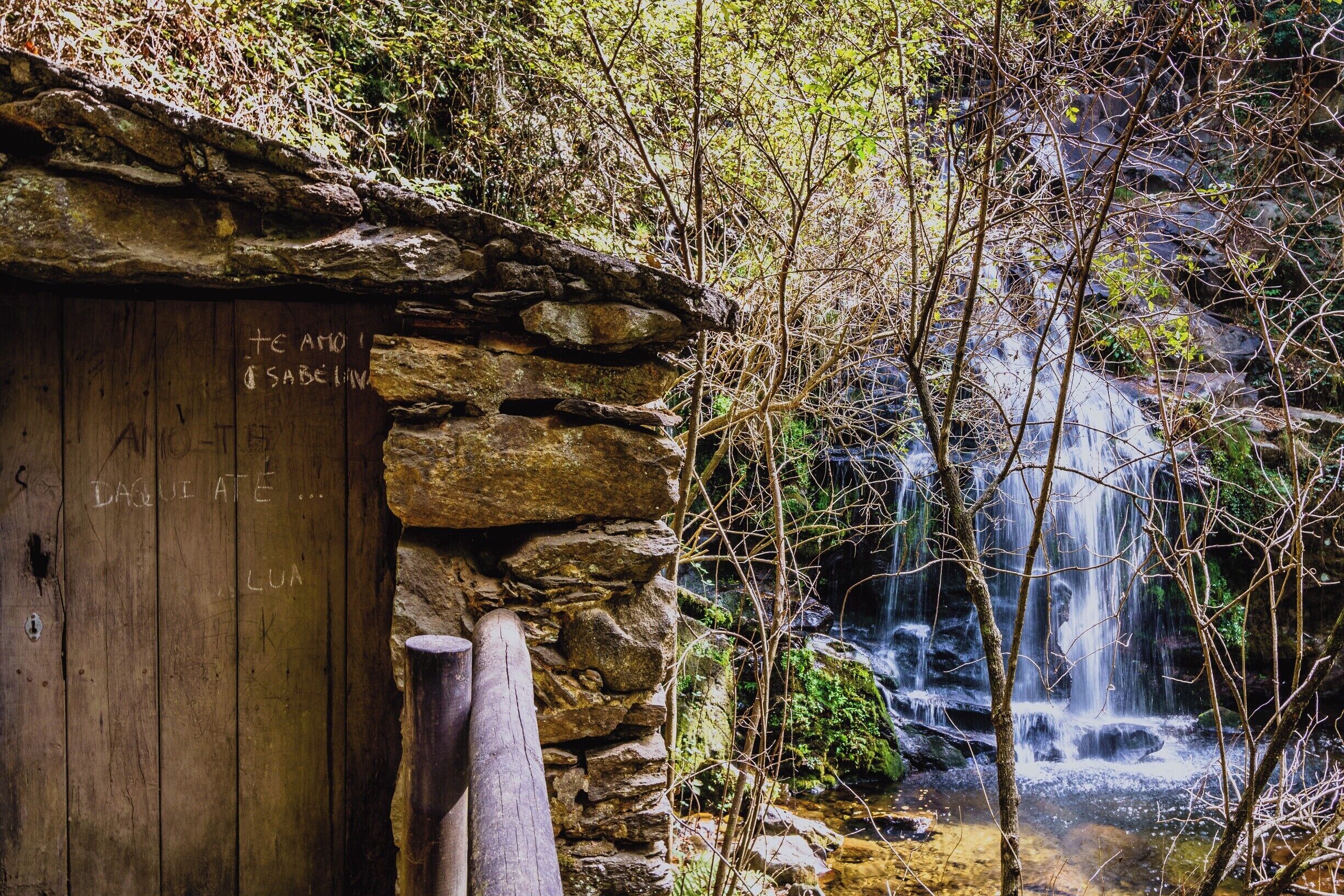 A stone building with graffiti, a wooden door, and a waterfall in the background.