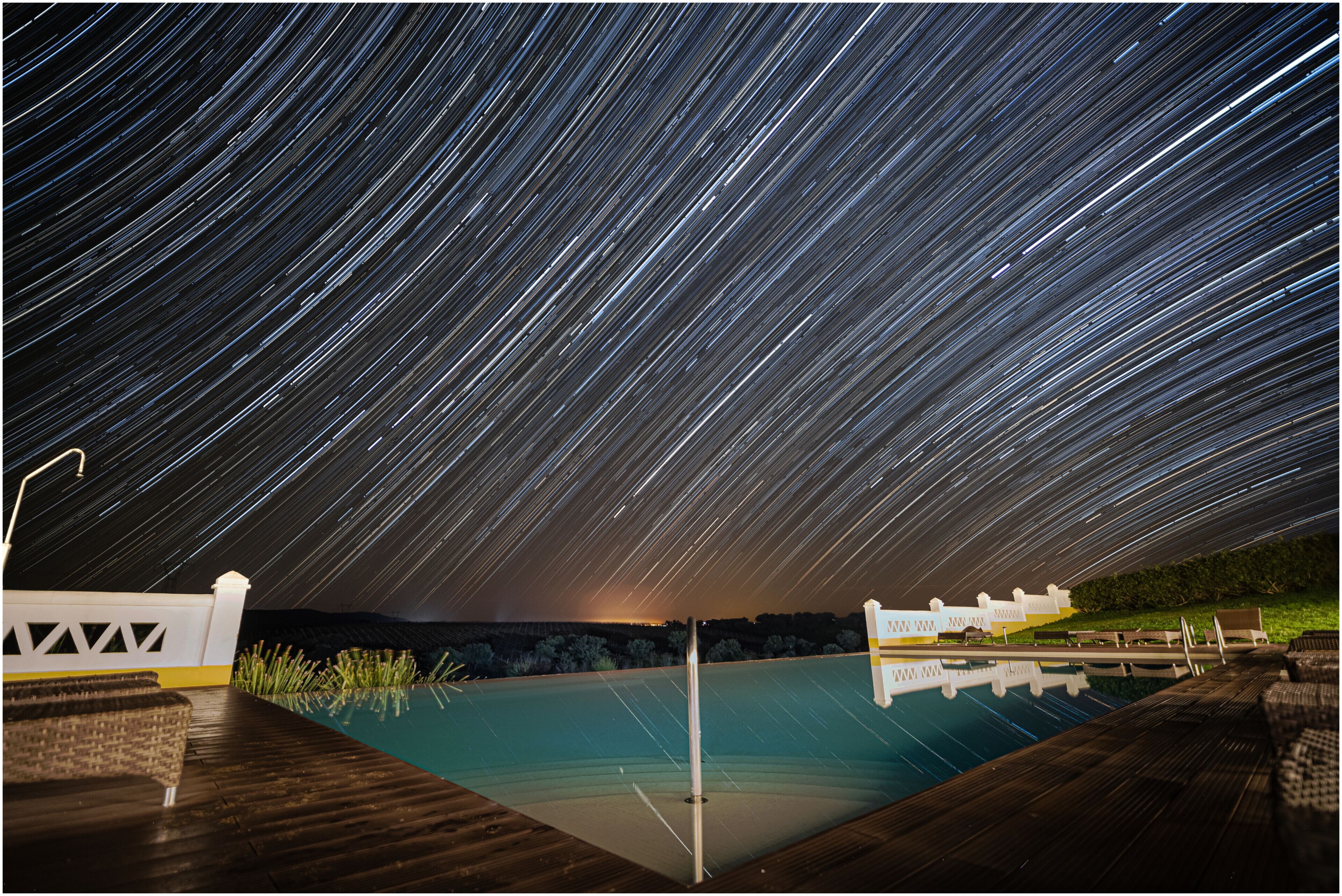 A starry night sky over a swimming pool area with wooden decking and lounge chairs.