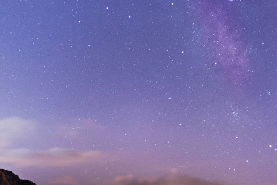 A starry night sky over a coastal landscape with a mountain in the foreground and a sunset in the background.