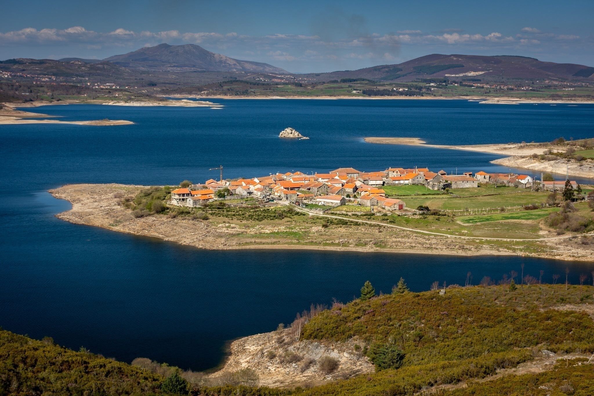 A coastal village with red-roofed buildings, a small island, and mountains in the background.