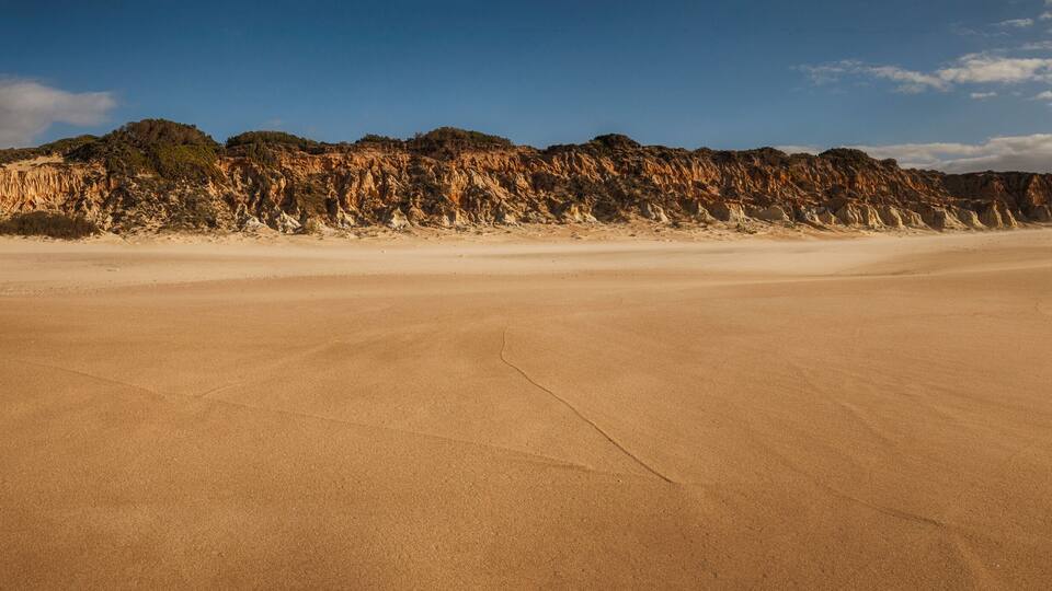A vast desert landscape with a prominent sand dune in the foreground and a rocky cliff in the background.