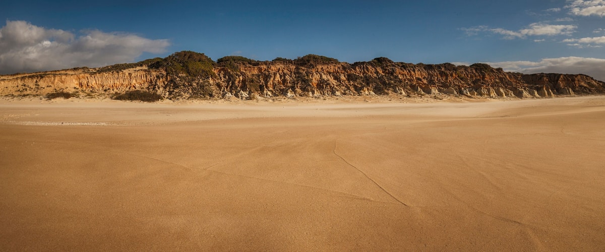 A vast desert landscape with a prominent sand dune in the foreground and a rocky cliff in the background.