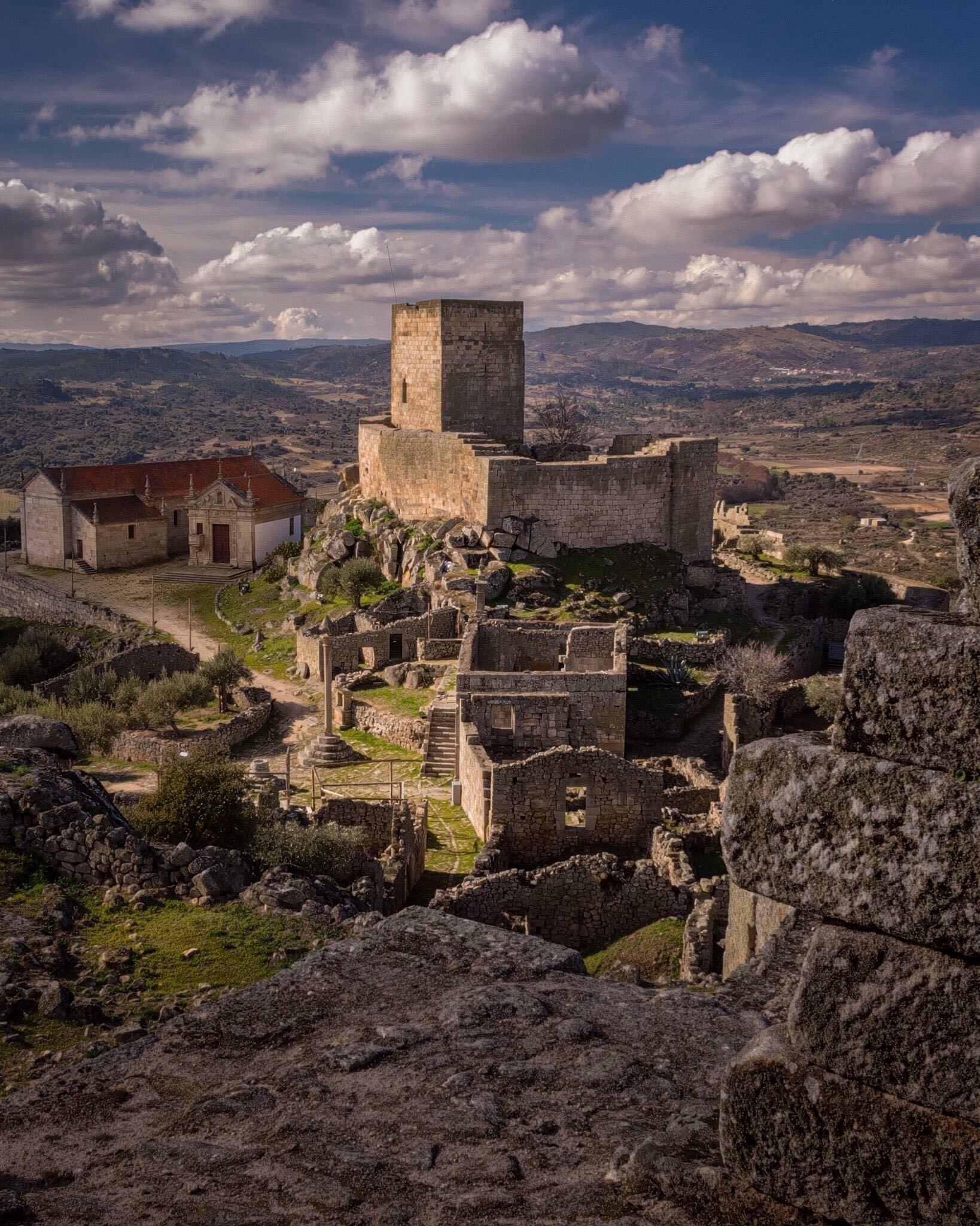 An ancient stone fortress with a prominent tower, surrounded by ruins and a small building with a red roof.