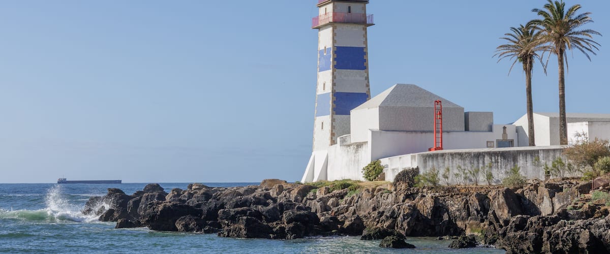 Un faro con la parte superior roja, un edificio blanco con una franja azul y palmeras junto al mar.