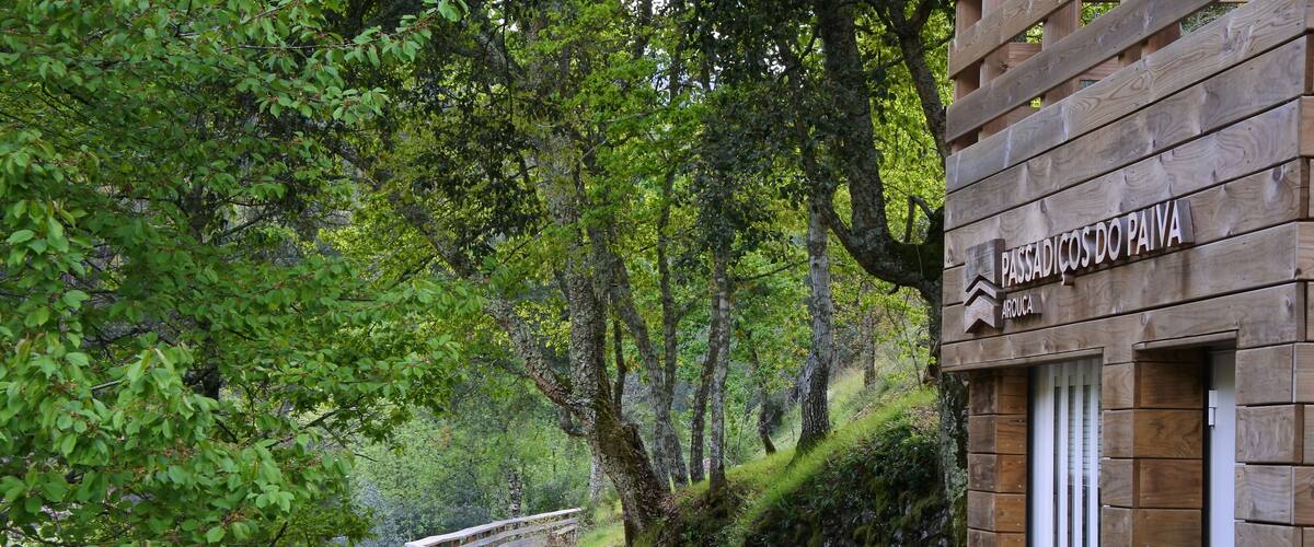 A wooden boardwalk leading through a forested area with a sign reading "PASSADICOS DO PAIVA."
