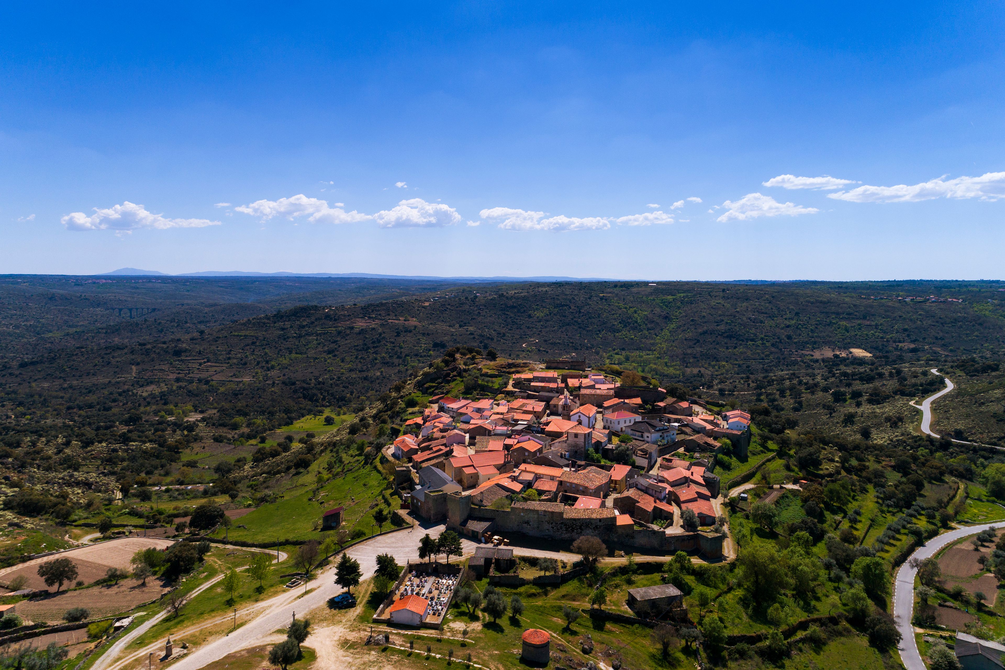 Vista aérea de un pueblo histórico con edificios de tejados rojos, rodeado de campos verdes y colinas.