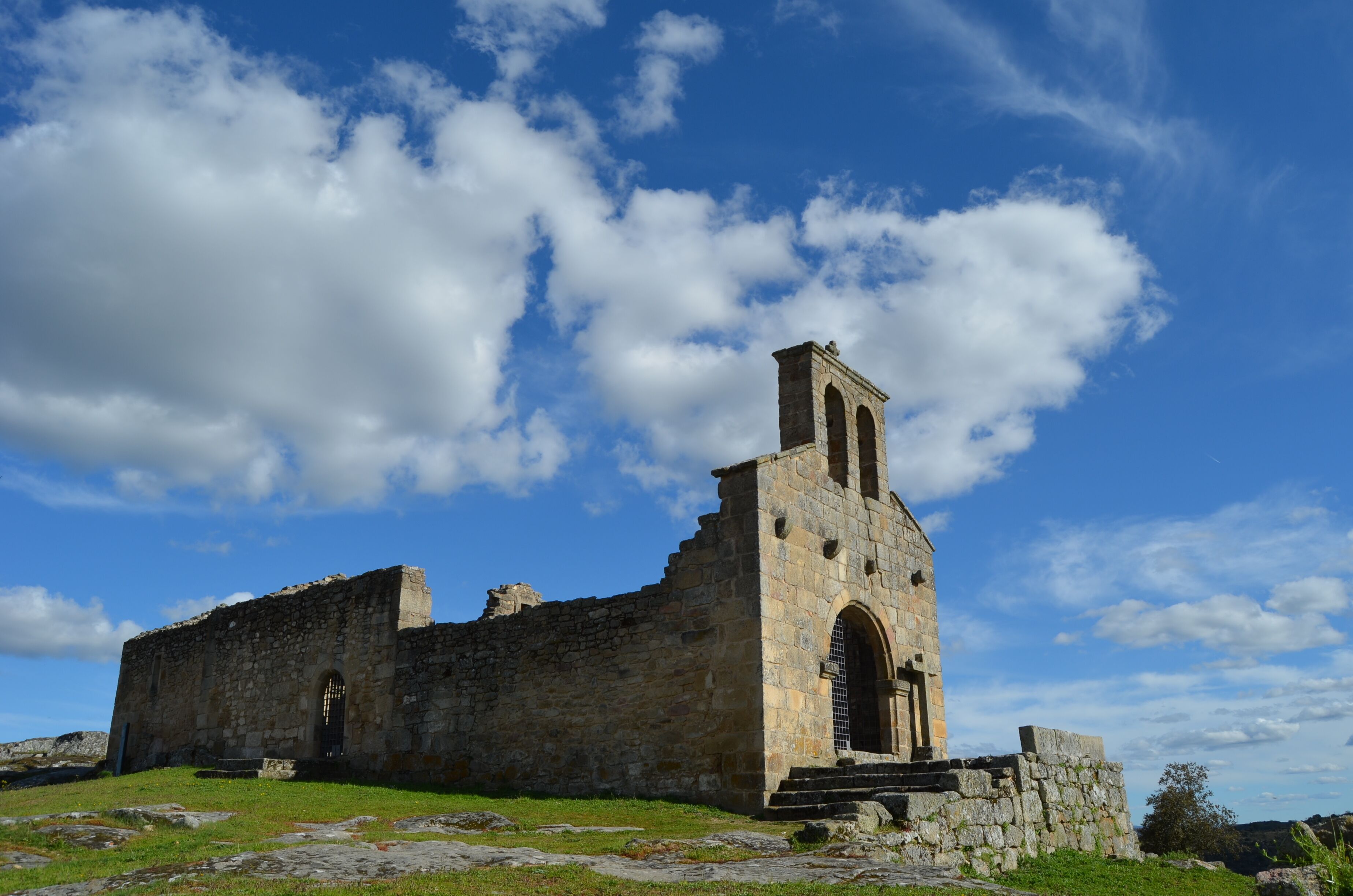 An ancient stone building with a bell tower, surrounded by grassy terrain and under a partly cloudy sky.