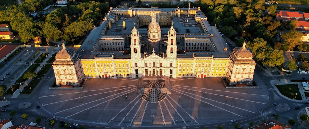 An aerial view of a grand building with two domes, surrounded by a spacious plaza and a dense forested area.