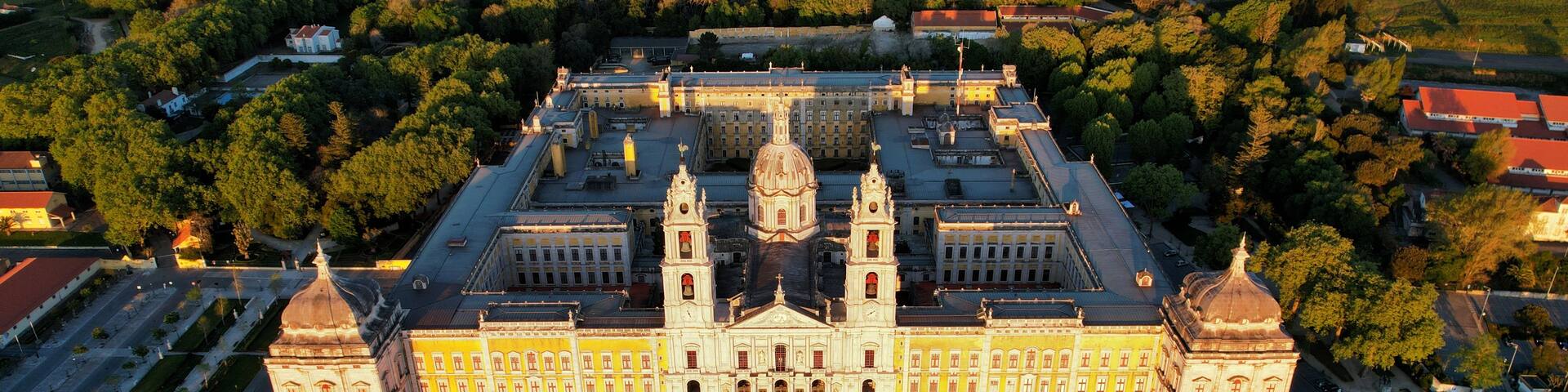 An aerial view of a grand building with two domes, surrounded by a spacious plaza and a dense forested area.