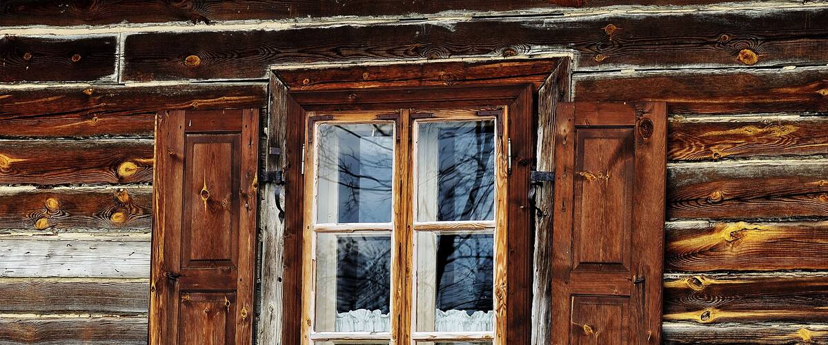 Una cabaña rústica de madera con una ventana que tiene dos persianas de madera y una cortina blanca.