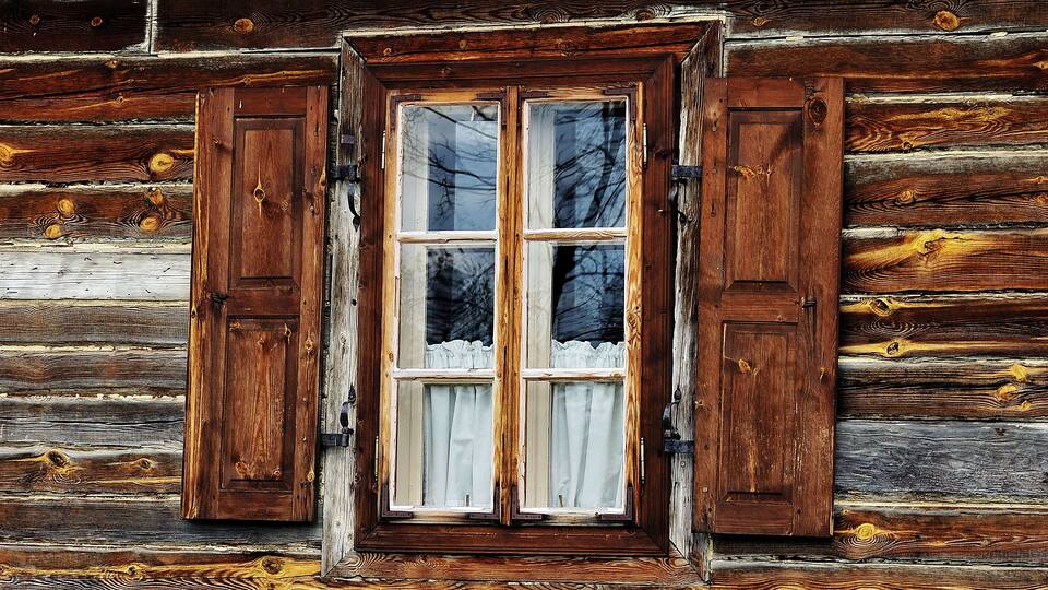 Eine rustikale Holzhütte mit einem Fenster, das zwei Holzklappen und einen weißen Vorhang hat.