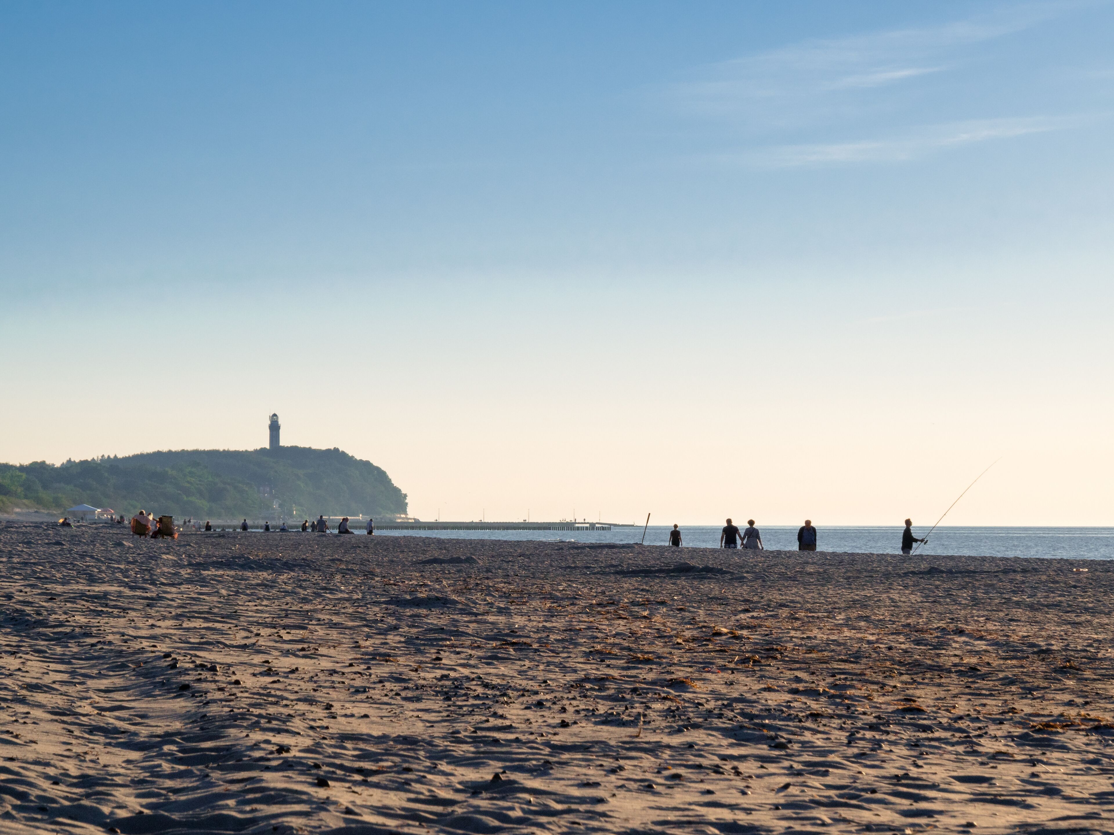 A beach with people fishing, a lighthouse, and a pier in the background.