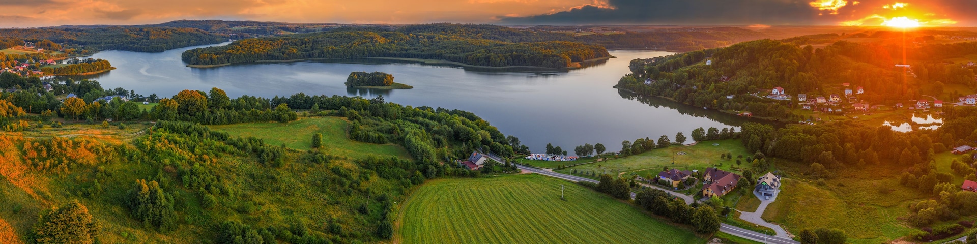 Um lago cercado por colinas arborizadas, uma pequena aldeia e um pôr do sol.
