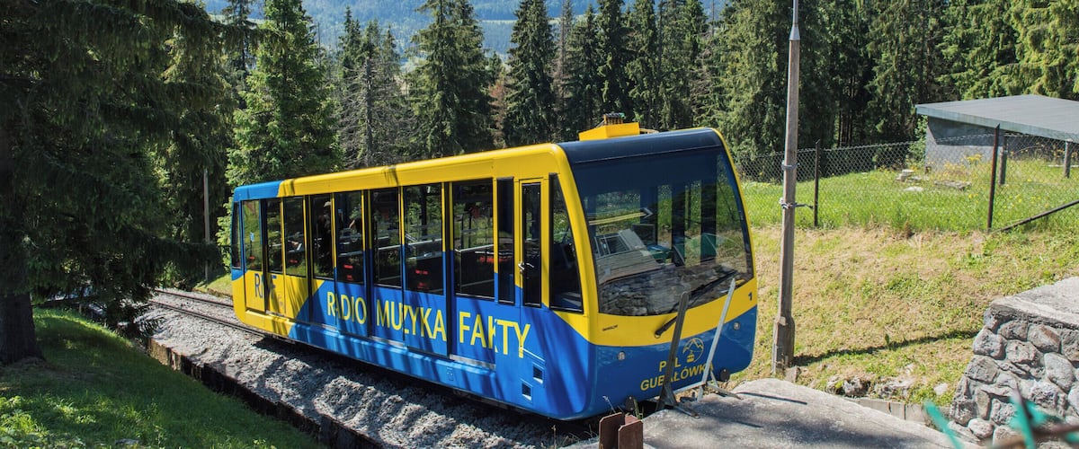 A blue and yellow cable car with "RDIO MUYKA FAHTY" written on it, traveling on tracks surrounded by trees and mountains.