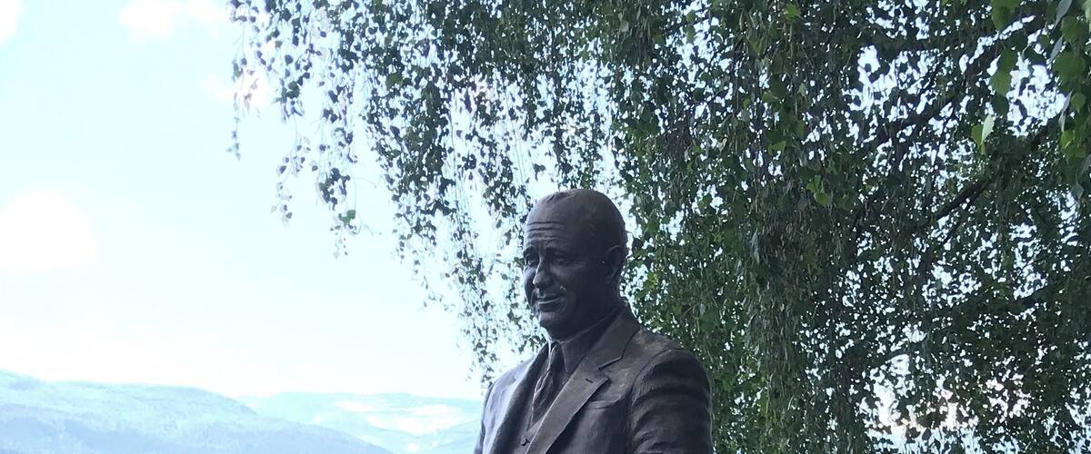 A statue of a man in a suit holding a football on a pedestal, with a lake and mountains in the background.