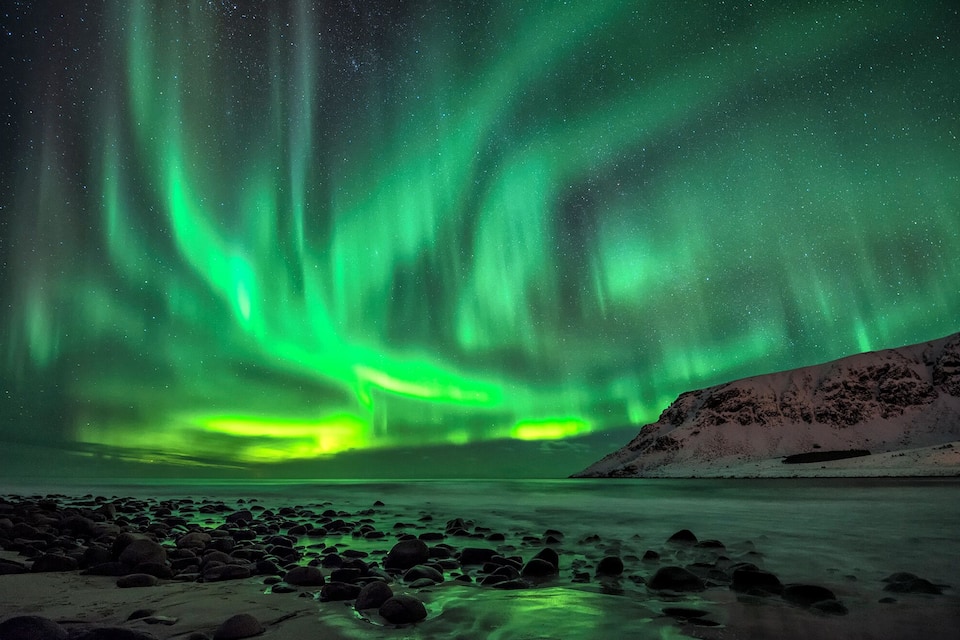The Northern Lights over a rocky shoreline with a snow-covered hill in the background.