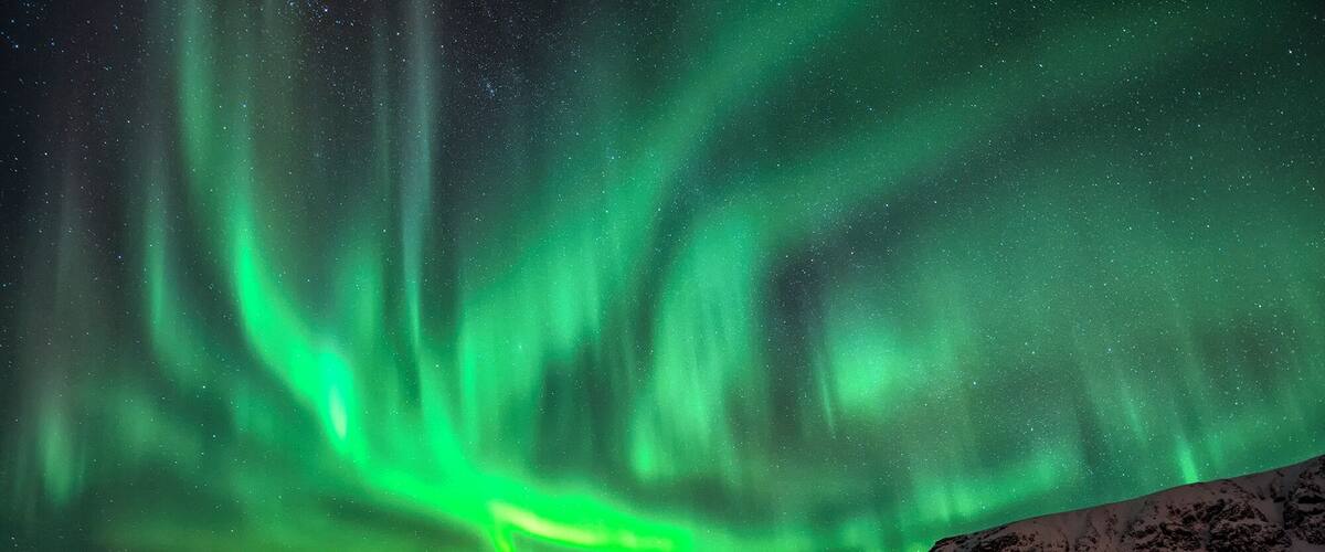 The Northern Lights over a rocky shoreline with a snow-covered hill in the background.