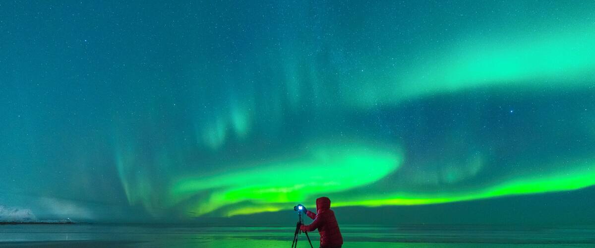 A person with a camera on a tripod, capturing the Northern Lights over a frozen landscape.