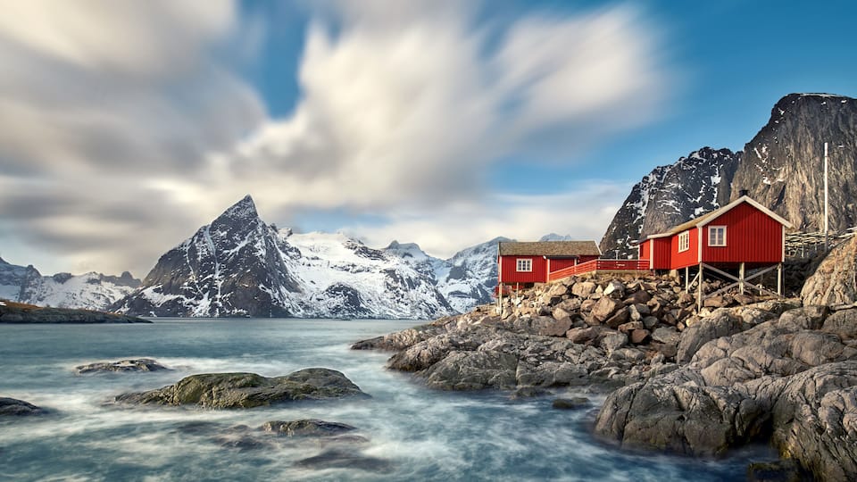 A red wooden house on a rocky shore with a mountain backdrop.