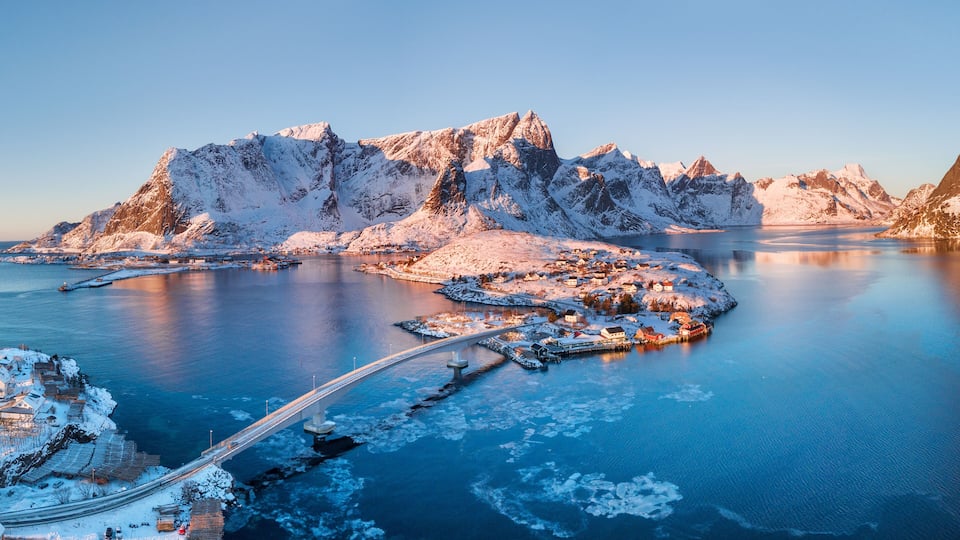 A snow-covered fjord with a bridge, mountains, and a small settlement.