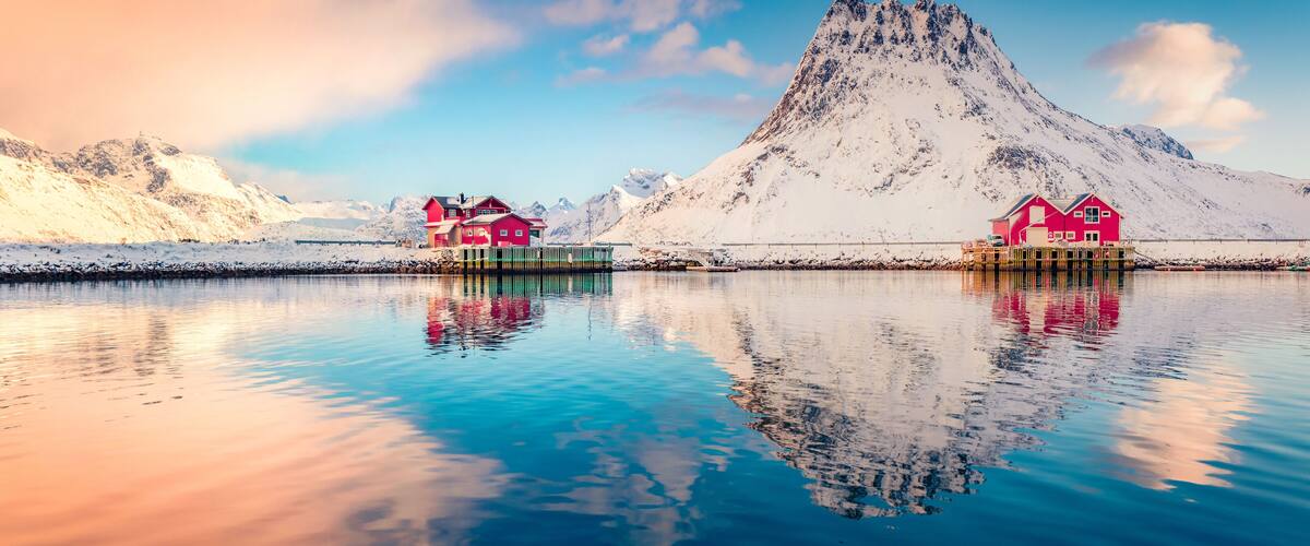 A snowy mountain with two red houses on stilts by a calm lake.