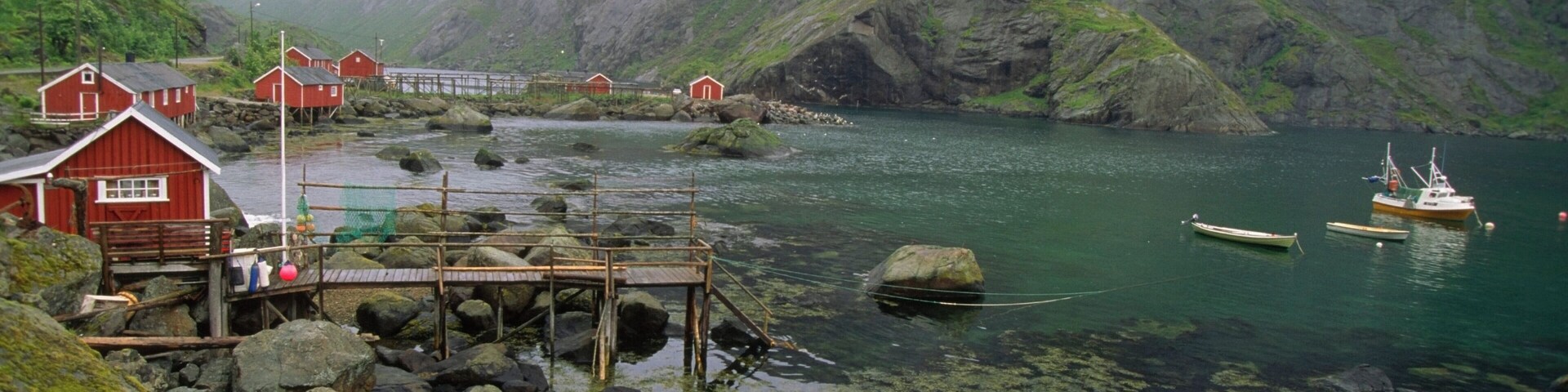 Uma área costeira rochosa com edifícios vermelhos, um cais de madeira e barcos num fiorde rodeado por penhascos íngremes.