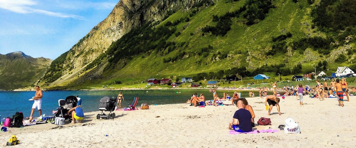 A beach with people sunbathing and swimming, surrounded by green hills and a clear blue sky.