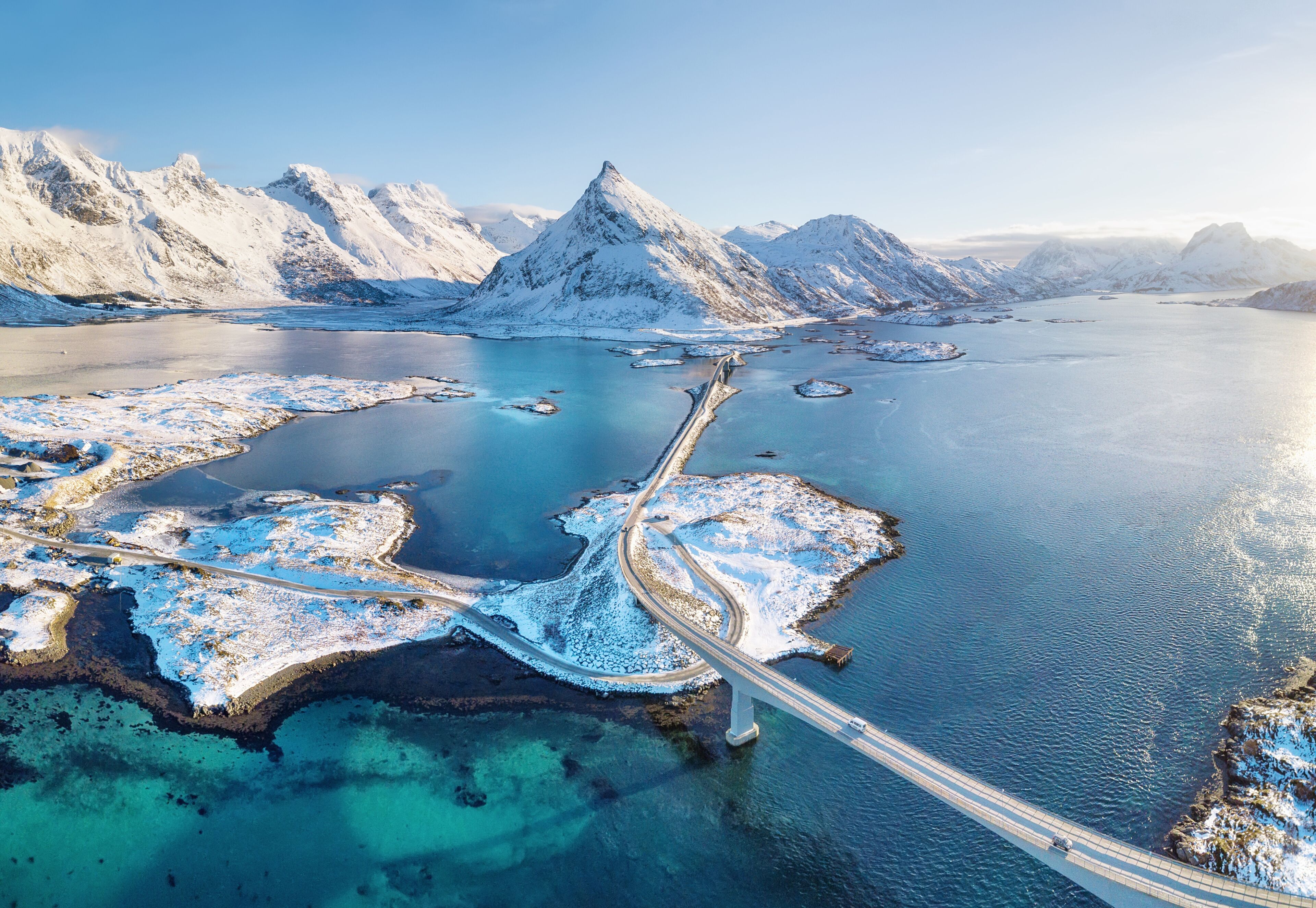 A snow-covered fjord with a bridge connecting islands, surrounded by mountains.