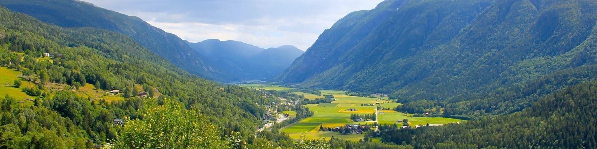 A valley with a river, surrounded by forested mountains and a few scattered buildings.