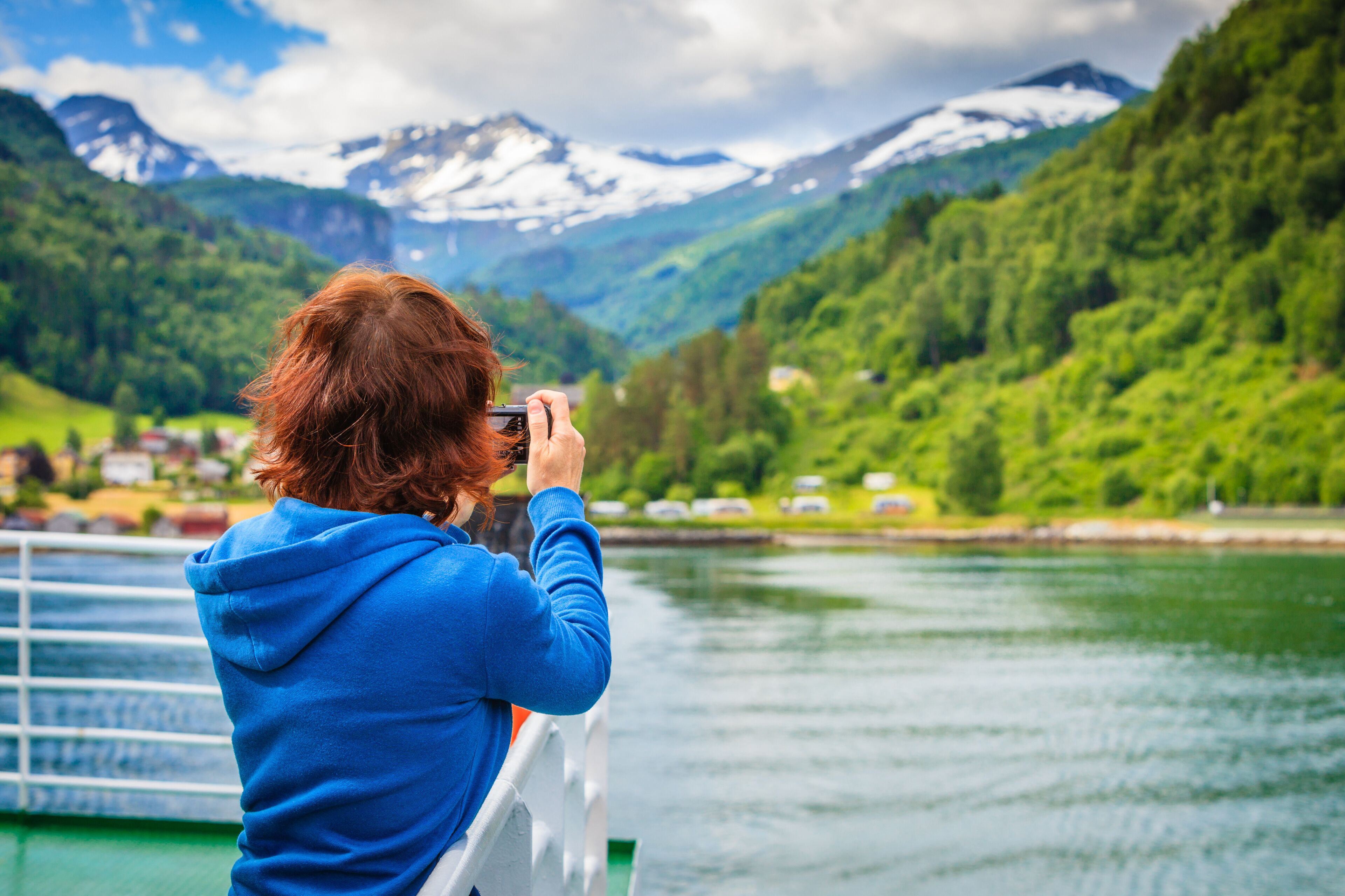 A person in a blue hoodie taking a photo of a lake with mountains in the background.