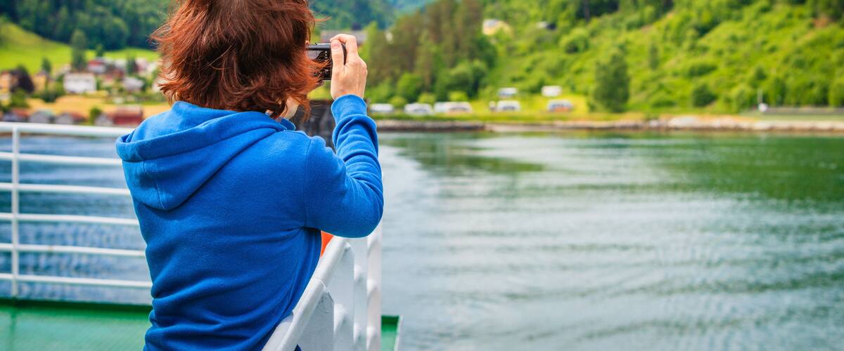 A person in a blue hoodie taking a photo of a lake with mountains in the background.